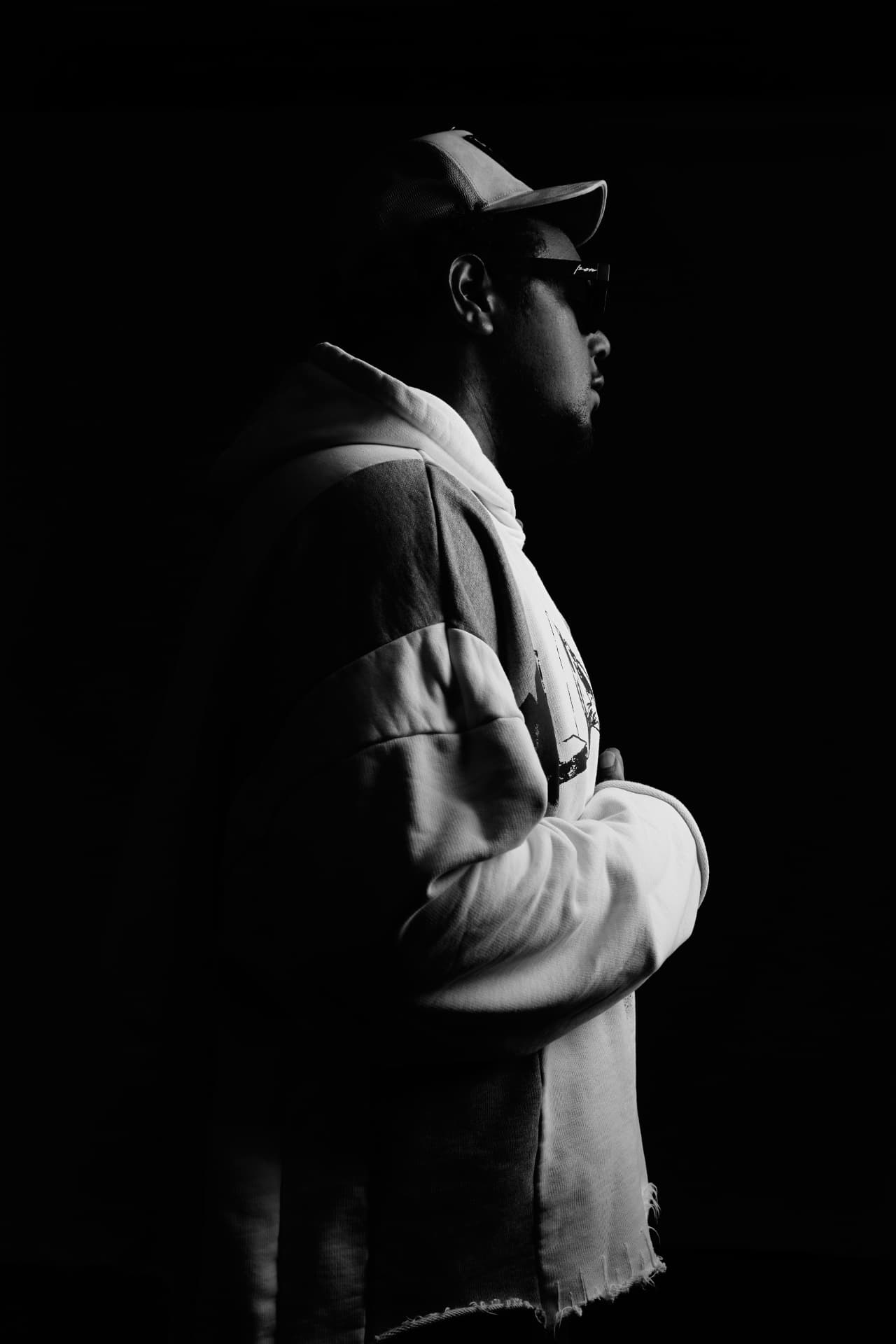 YO.G.I in profile, oversized layered hoodie and dark shades, lit from one side in deep shadow